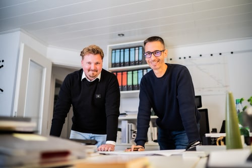 Zwei lächelnde Männer stehen in einem Büro über Bauplänen, Ordnerregal im Hintergrund.