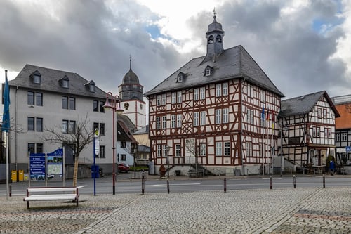 Historisches Rathaus mit Fachwerkfassade und Turm, umgeben von Gebäuden und einem gepflasterten Platz unter bewölktem Himmel.