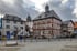 Historisches Rathaus mit Fachwerkfassade und Turm, umgeben von Gebäuden und einem gepflasterten Platz unter bewölktem Himmel.
