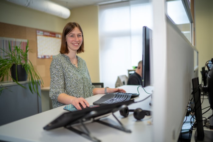 Eine lächelnde junge Frau mit braunen Haaren sitzt an einem Schreibtisch mit zwei Monitoren und einer Pflanze im Büro.