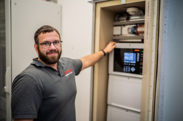Mann mit Brille und Bart zeigt auf ein installiertes System mit Bildschirm und Bedienelementen in einem Schrank.