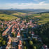 Luftaufnahme eines Dorfes mit roten Dächern, Kirchturm, Fluss, Feldern und Bergen unter blauem Himmel.