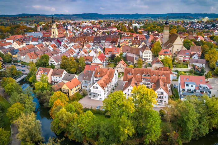 Luftbild einer deutschen Stadt mit roten Dächern, Kirchtürmen und einem Fluss, der durch grüne Bäume fließt.
