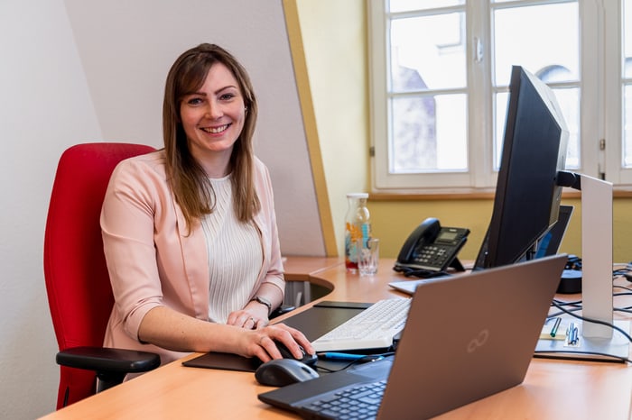 Frau mit langen braunen Haaren, rosa Blazer und weißem Oberteil lächelt am Schreibtisch mit Laptop und zwei Monitoren.