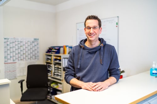 Zwei lächelnde Männer stehen in einem Büro über Bauplänen, Ordnerregal im Hintergrund.