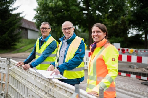 Drei Bauarbeiter in leuchtenden Westen lächeln vor einer Baustelle mit Absperrungen und Lichtern.