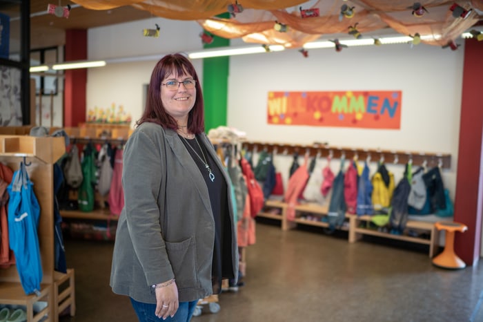 Eine lächelnde Frau steht in einem Kindergartenflur mit Kinderkleidung und einem "WILLKOMMEN"-Banner.