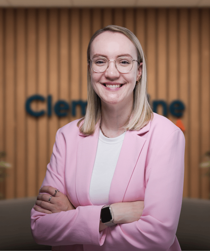 Lachende blonde Frau mit Brille und rosa Blazer, Arme verschränkt, vor Holzwand mit "Clementine"-Logo.