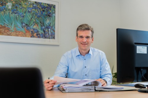 Laechelnder Mann mit Brille im hellblauen Hemd sitzt an seinem Schreibtisch und blaettert in Unterlagen, im Hintergrund ein Bildschirm und ein gerahmter Kunstdruck.