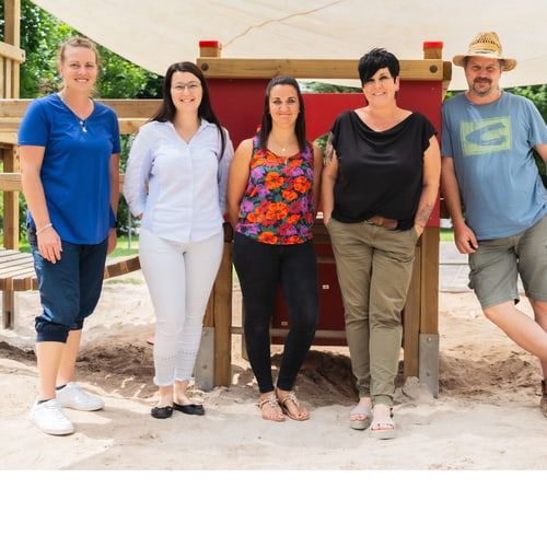 Fünf Erwachsene stehen lächelnd vor einem Spielplatz mit Sand und rotem Spielgerät.