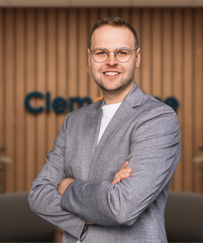 Ein lächelnder Mann mit Brille und grauem Sakko, die Arme verschränkt, vor einer Holzwand mit "Clem" Logo.