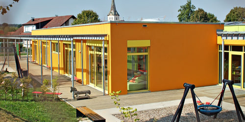 Helles orangefarbenes Kindergartengebäude mit vielen Fenstern, Spielplatz mit Schaukeln und Holzpferd.