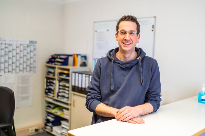 Ein lächelnder Mann mit Brille und blauem Kapuzenpullover lehnt an einem weißen Schreibtisch in einem Büro.