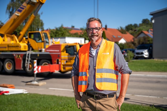 Mann mit Brille und orangefarbener Warnweste vor gelbem Kran und Straße.