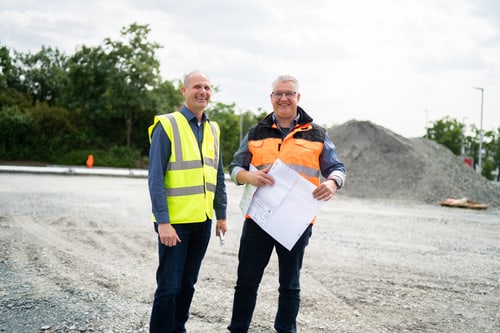 Zwei lächelnde Bauarbeiter in Warnwesten stehen auf einer Baustelle mit Kies und Bauplänen.