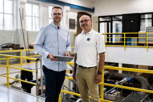Zwei Männer im Wasserwerk des Zweckverbands RiesWasserVersorgung stehen auf einer Empore mit gelbem Geländer, einer von ihnen hält ein Tablet.
