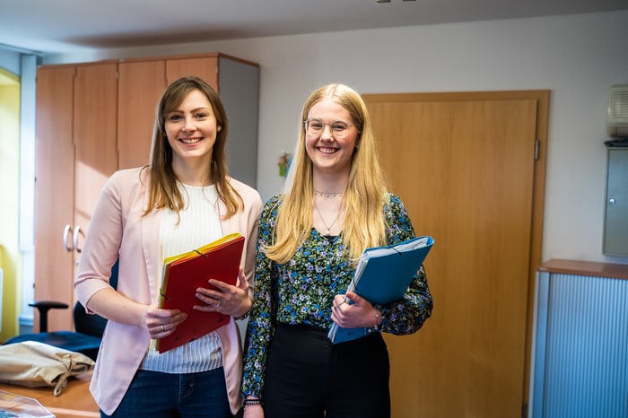 Zwei lächelnde junge Frauen halten Ordner in einem Büro.