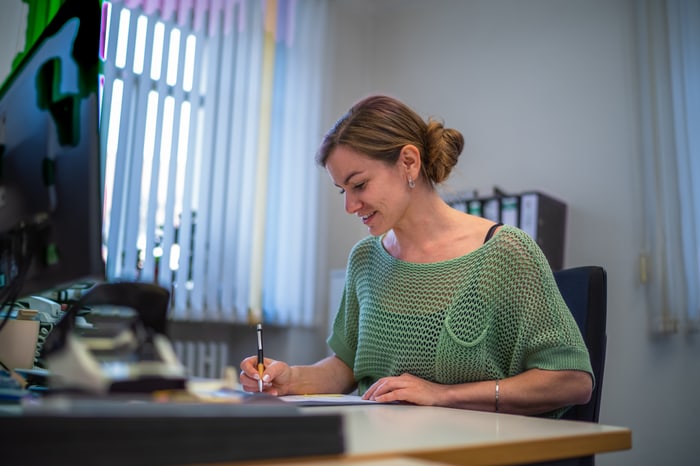 Junge Frau in grünem Pullover sitzt lächelnd am Schreibtisch und schreibt mit einem Stift.