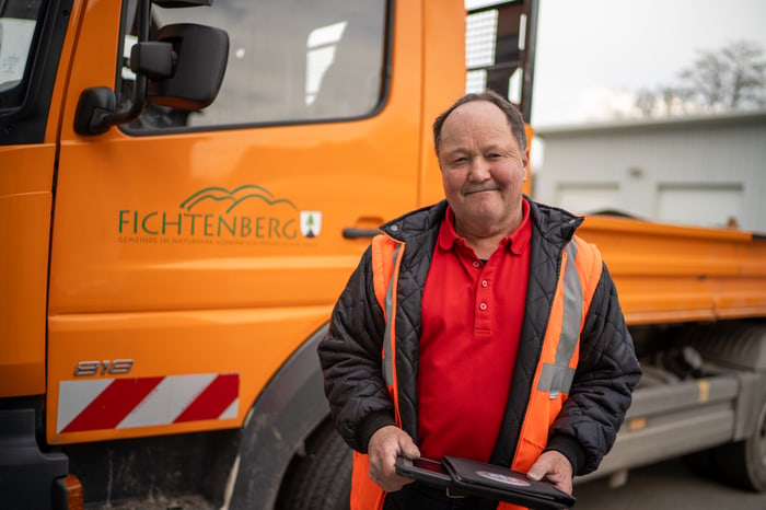 Mann in Warnweste vor orangefarbenem LKW mit "FICHTENBERG" Logo und Tablet in der Hand.