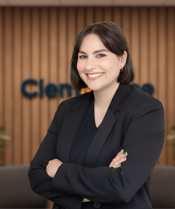 Lächelnde Frau mit kurzen, dunklen Haaren und schwarzem Blazer vor einer Holzwand mit „Clemence“-Logo.