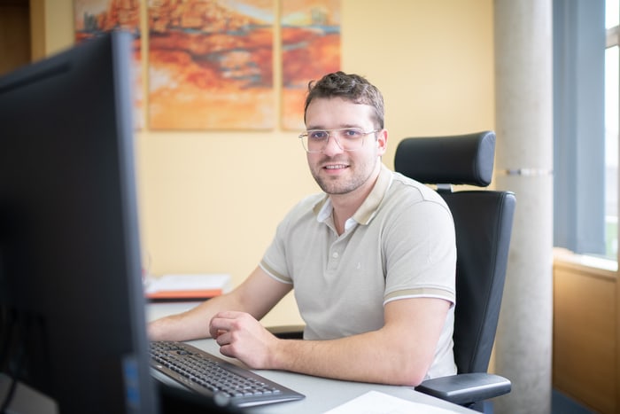Mann mit Brille sitzt am Schreibtisch vor einem Computerbildschirm und lächelt in die Kamera.