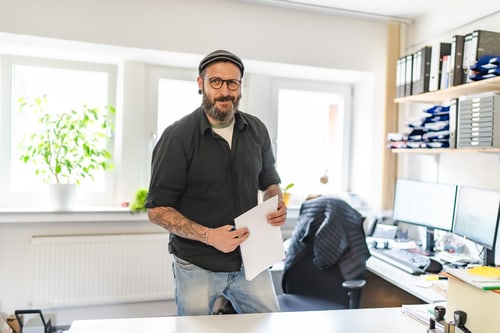 Ein lächelnder Mann mit Bart, Mütze und Brille hält Papiere in einem sonnigen Büro.